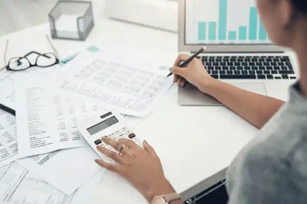 Woman auditing papers with calculator, pen, and MacBook